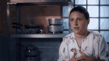 a woman in a floral shirt is talking in front of a shelf with pots and pans .