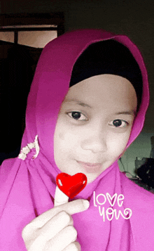 a girl wearing a pink hijab holds a red heart shaped lollipop