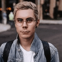 a young man wearing glasses and a denim jacket is standing in front of a building .