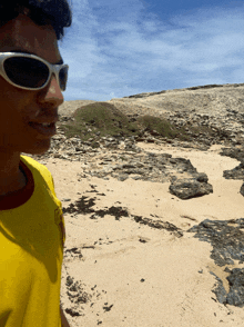 a man wearing sunglasses and a yellow shirt stands on a rocky beach