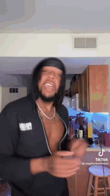 a shirtless man is standing in a kitchen wearing a black jacket and a necklace .