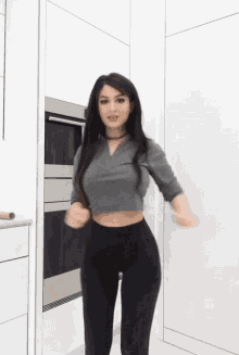 a woman in a grey crop top and black leggings is dancing in a kitchen