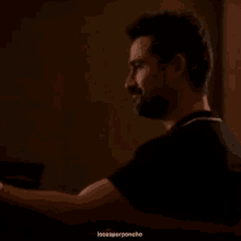 a man with a beard is sitting at a table in a dark room looking at something .