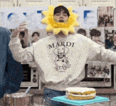 a person wearing a sun hat and a mardi sweatshirt is standing in front of a cake on a table .