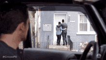a man is driving a car and looking out the window at a couple standing in front of a house .