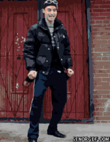a man in a black jacket stands in front of a red door with senorgif.com written on the bottom