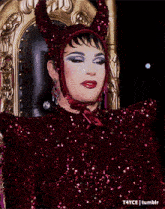 a drag queen is sitting on a throne wearing a red sequined dress and horns .