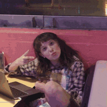 a girl in a plaid shirt is sitting in front of a laptop and making a peace sign