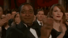 a man in a tuxedo is applauding while a woman in a dress looks on .