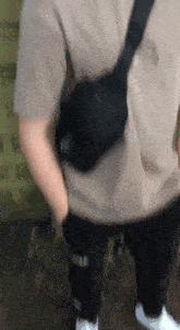 a man wearing a tan shirt and black pants is standing with his hands in his pockets and a black bag on his shoulder .