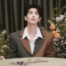 a man in a suit and white shirt is sitting at a table with flowers .
