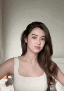 a woman in a white tank top and necklace is standing in front of a white wall .