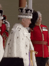 a man wearing a crown and a fur coat is standing in front of a group of people in uniform .