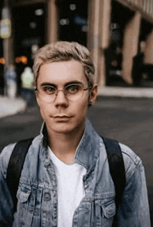 a young man wearing glasses and a denim jacket is walking down a street
