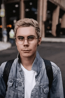 a young man wearing glasses and a denim jacket stands in front of a building