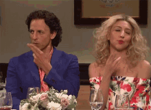 a man in a blue suit and a woman in a floral dress are sitting at a table with wine glasses and flowers .