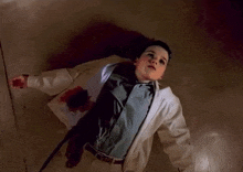 a boy in a white jacket is laying on the floor with blood on his shirt