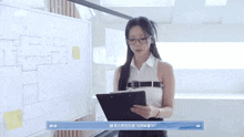 a woman in glasses is holding a clipboard in front of a whiteboard .