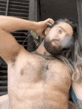 a shirtless man with a beard and long hair holds his hand to his head