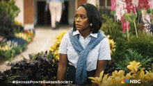 a woman is sitting in a garden with a nbc logo in the background