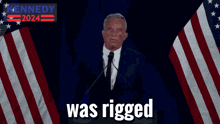 a man in a suit and tie is giving a speech in front of an american flag and says kennedy 2024