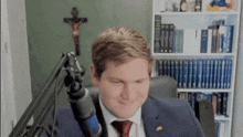 a man in a suit and tie is sitting in front of a microphone in a room with a cross in the background .
