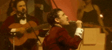 a man in a red suit is singing into a microphone while another man plays guitar .