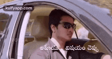 a man wearing sunglasses is driving a car with a caption in telugu .
