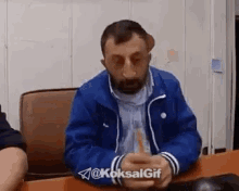 a man with a beard and a blue jacket is sitting at a table with his hands folded .