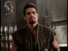 a man with a beard and a leather vest is standing in a room .