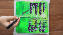 a person is holding a spatula over a painting of purple flowers on a green background