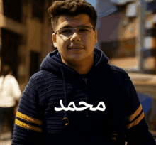a young man wearing glasses and a blue sweater has the name muhammad written on his shirt