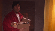 a man in a red jacket is holding a cardboard box and a can of soda