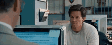 a man is sitting at a desk in front of a computer and looking at another man .