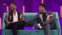 two men are sitting on a blue couch holding hands and laughing with the letters rtve visible in the background