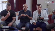a group of men are sitting in a circle eating food with a tv logo in the background .