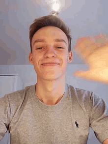 a young man wearing a grey polo shirt is smiling and waving at the camera