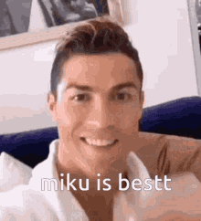 a man is sitting on a couch and smiling with the words miku is bestt written below him .