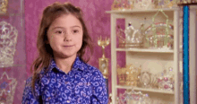 a little girl in a blue shirt is standing in front of a shelf filled with tiaras .