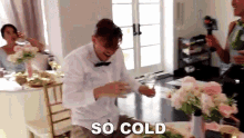 a man in a bow tie is sitting at a table with flowers and drinking from a glass with the words so cold above him .