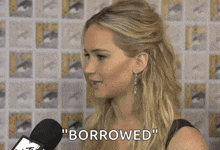 a woman is talking into a microphone and says " borrowed " in front of a comic con wall