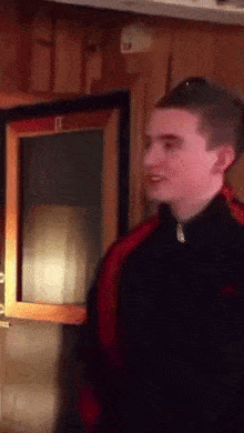 a young man in a black and red jacket is standing in front of a window