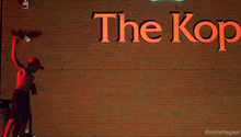 a man stands in front of a brick wall with a sign that says the kop