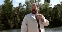 a bald man with a beard is wearing a white coat and tie and standing in front of a forest .