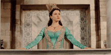 a woman in a green and gold dress with a crown on her head stands on a balcony