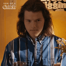 a man with long hair is wearing a blue and white striped shirt with the words son of a grifch behind him