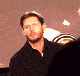 a man with a beard wearing a black shirt is standing in front of a screen .
