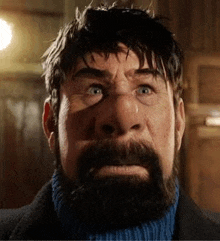 a man with a beard and mustache is wearing a blue turtleneck sweater and looking at the camera .