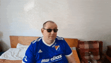 a man wearing sunglasses and a blue adidas shirt sitting on a bed