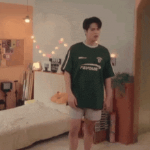 a young man in a green shirt and shorts is standing in a bedroom next to a bed .